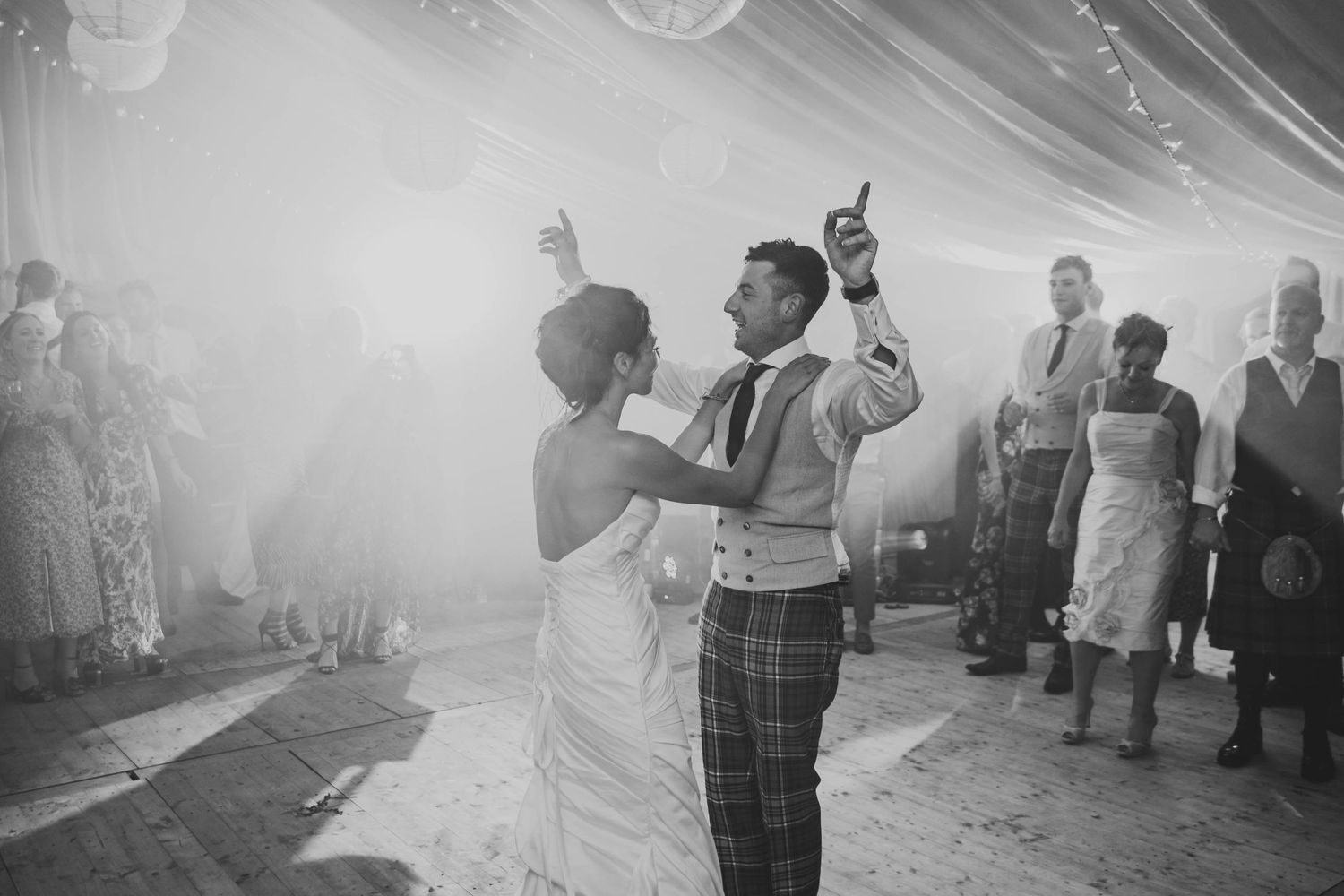 A black and white photo captures newlyweds sharing their first dance under atmospheric lighting.