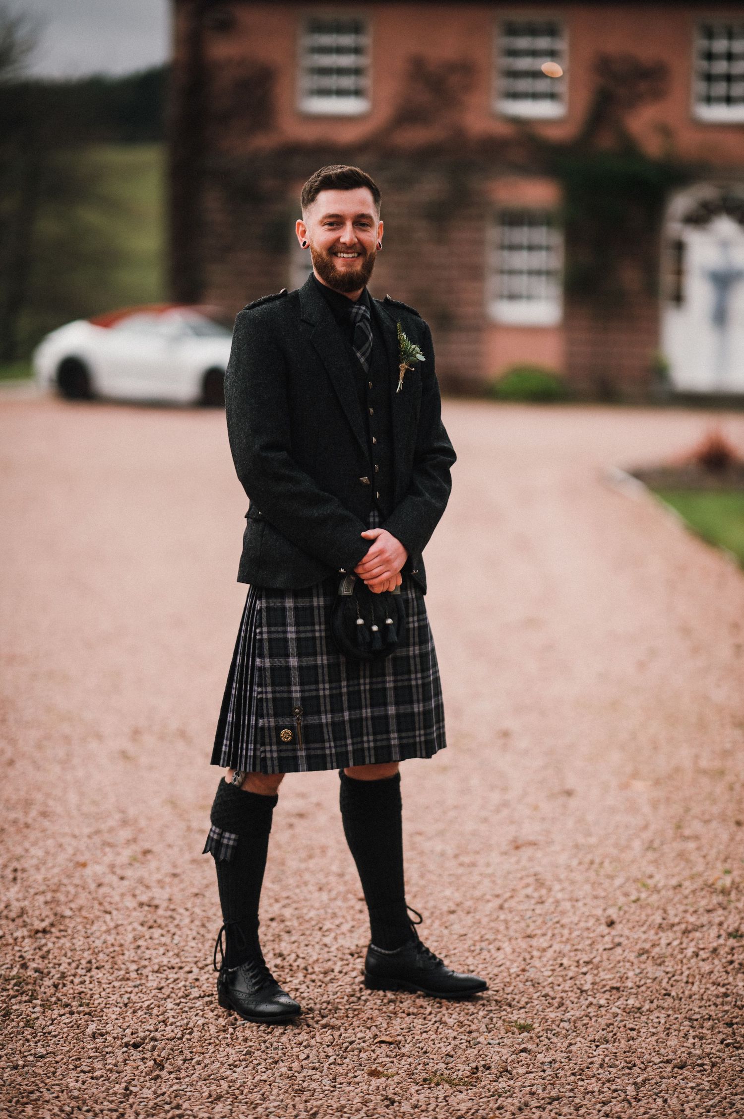 Person wearing a black jacket and traditional Scottish kilt stands on gravel driveway with brick building in background.