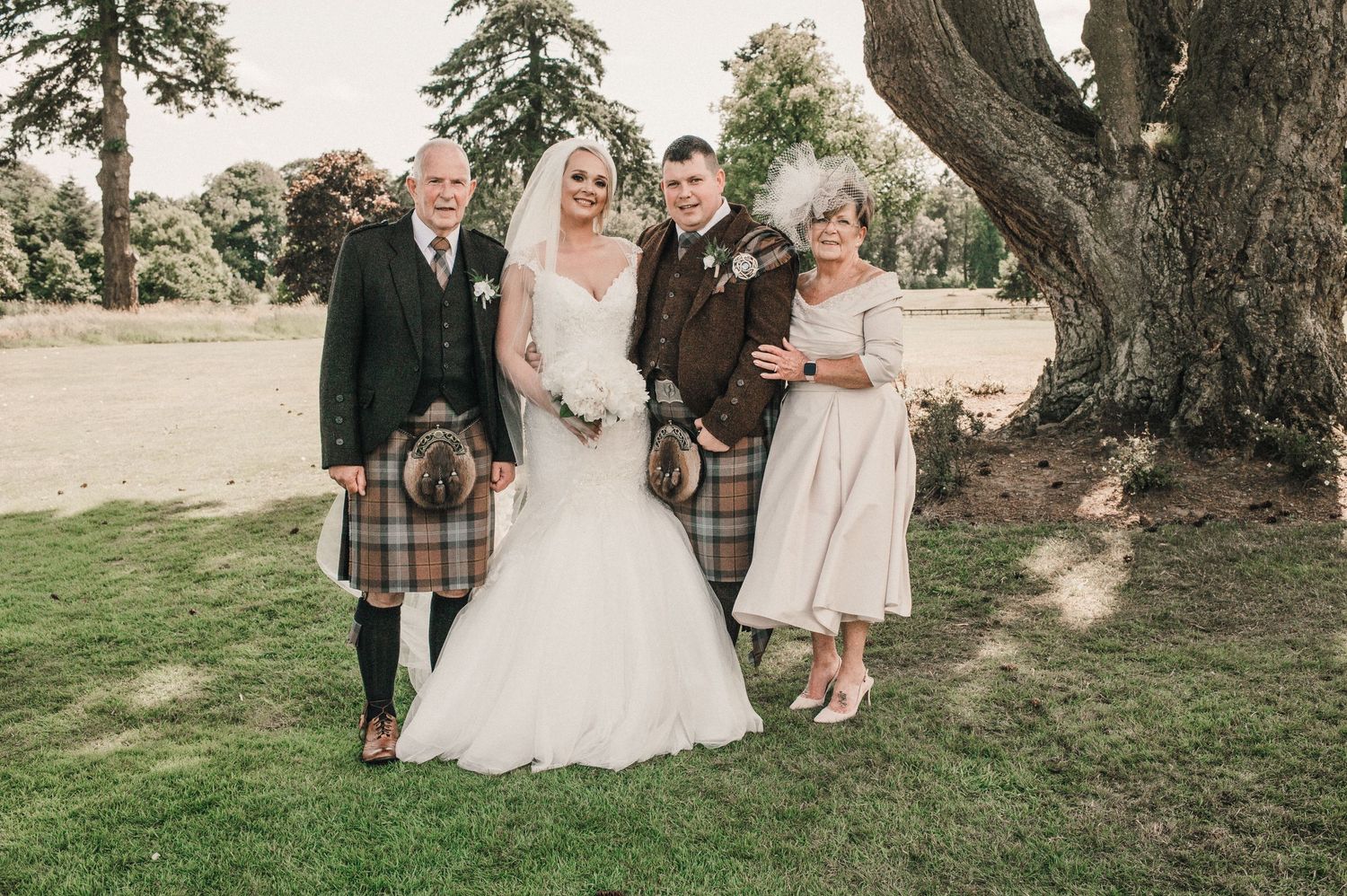 Four bridesmaids in cream-colored dresses pose with bouquets under a large tree.