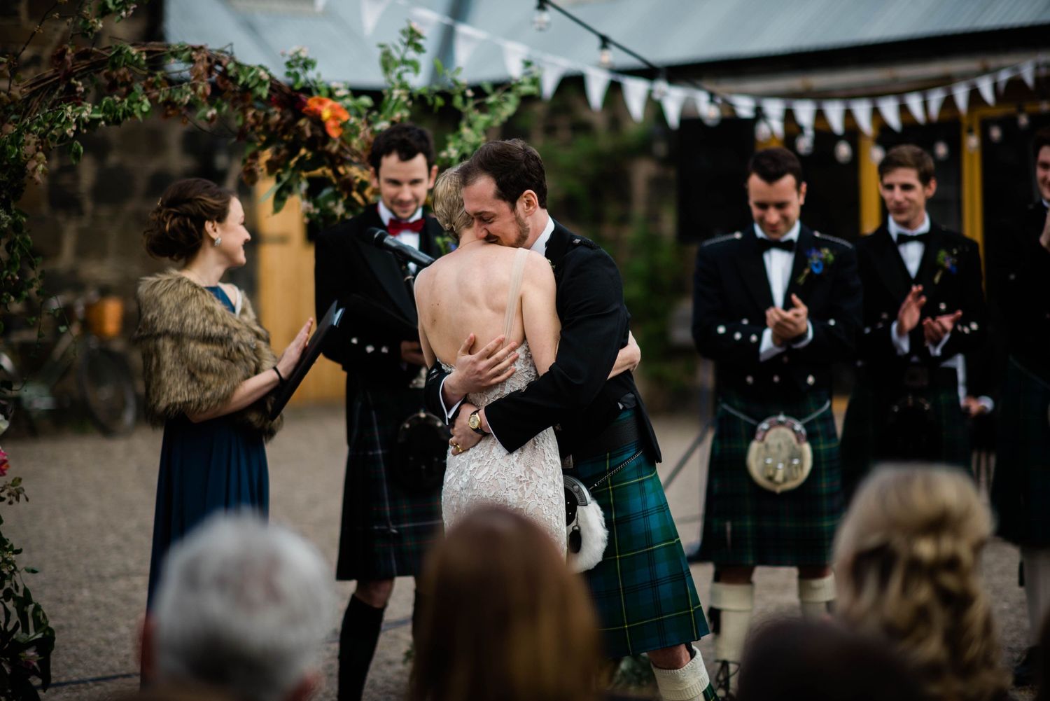 A couple celebrates their Scottish wedding ceremony outdoors under string lights with guests in kilts watching.