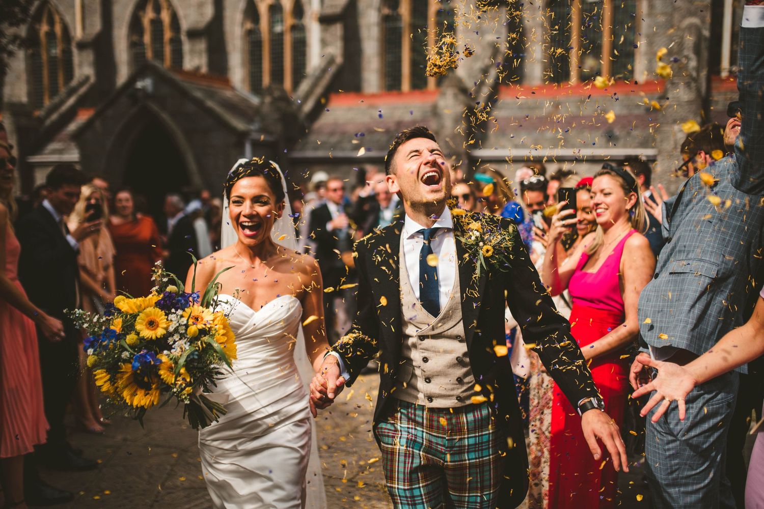 Jubilant wedding celebration with confetti and sunflowers outside gothic church.