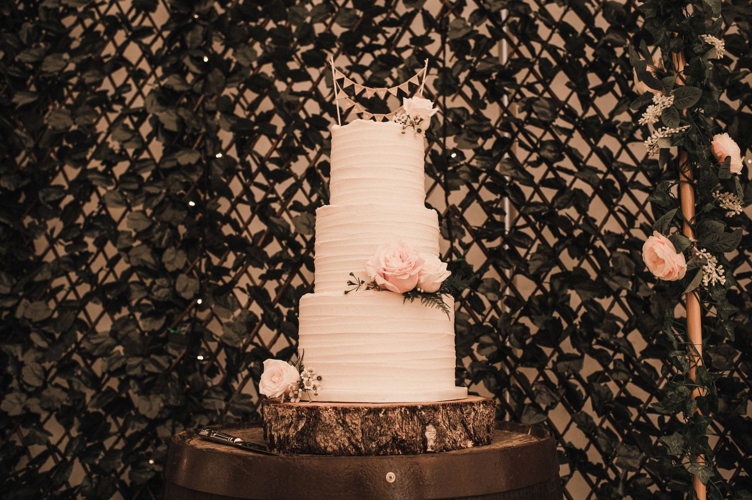 White tiered wedding cake with pink floral accents displayed against a dark lattice backdrop.