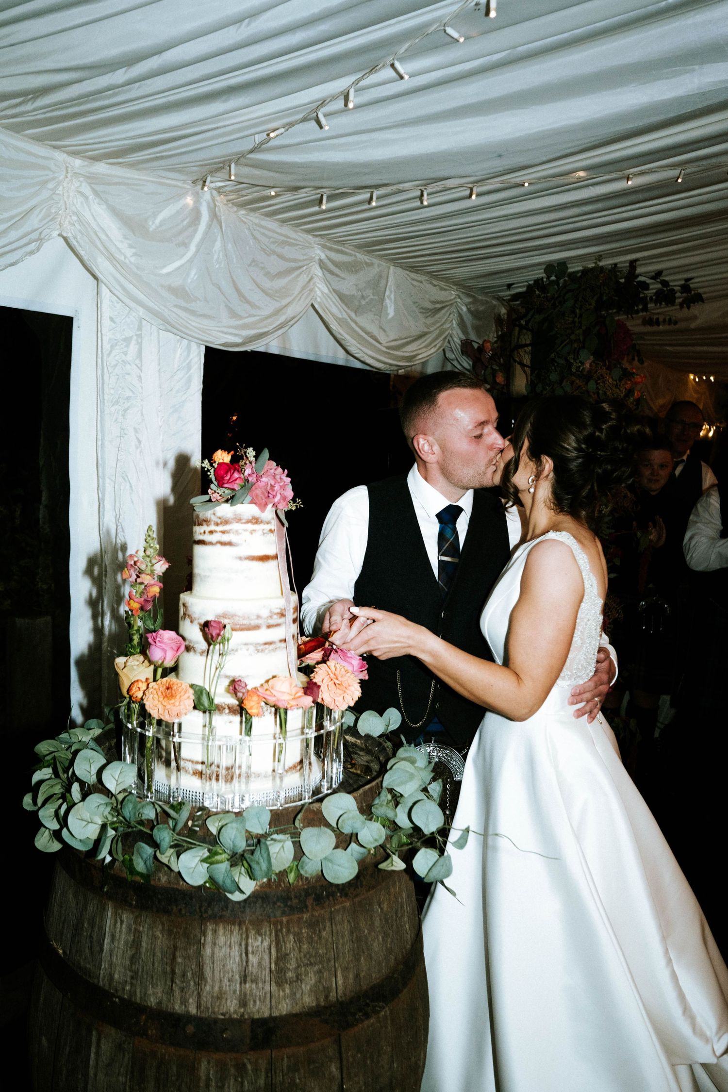 Wedding cake display with eucalyptus and pink flowers on rustic wooden stand at reception.
