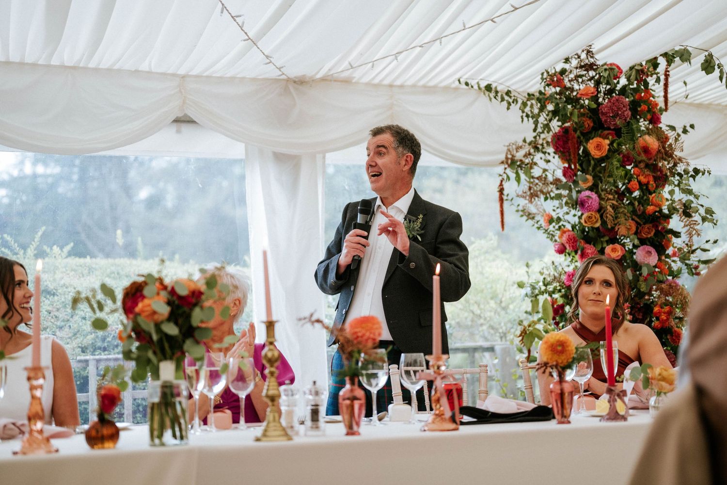 A speech being given at a wedding reception under a white marquee tent with floral decorations.