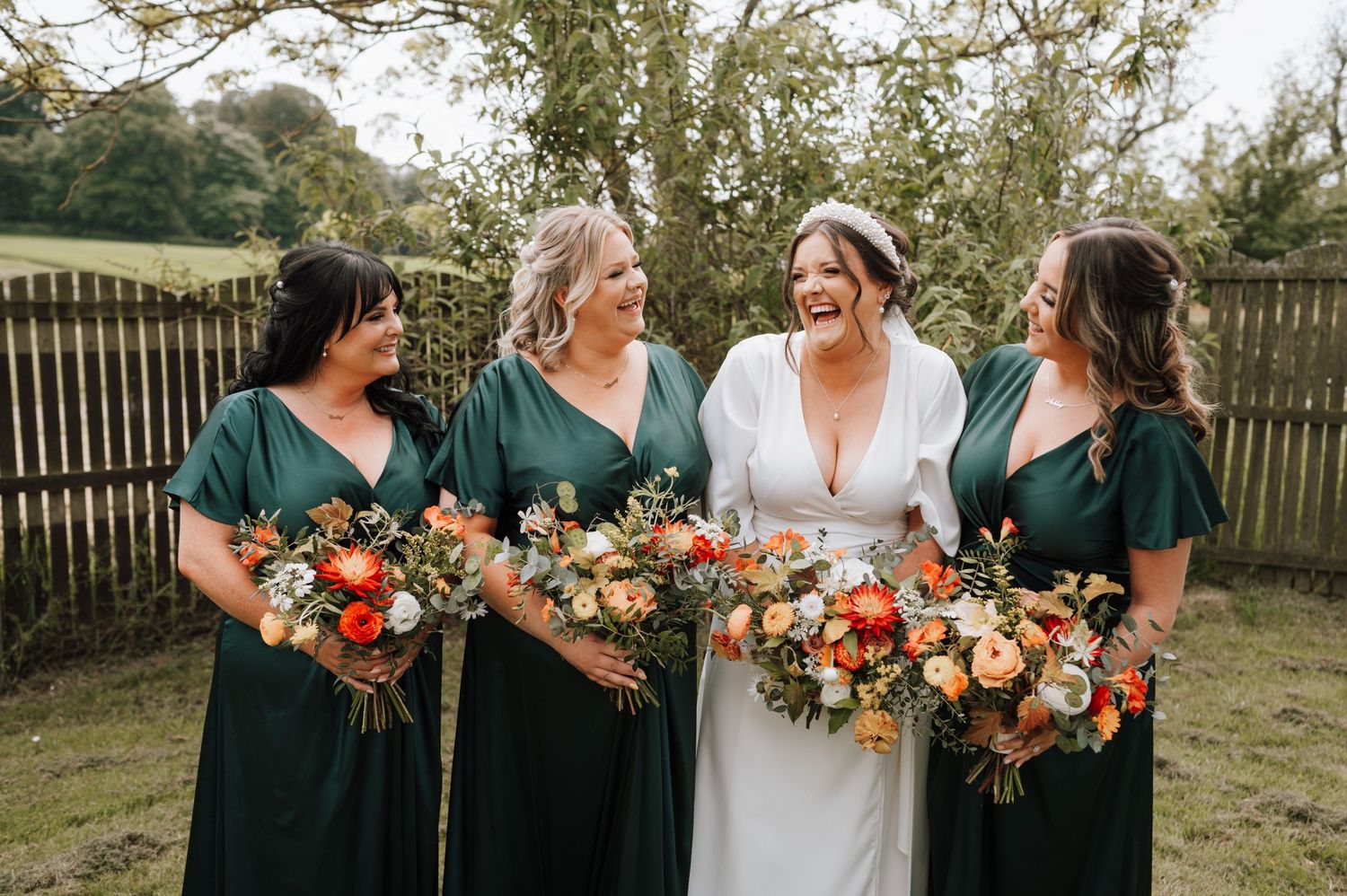 Bridal party poses outdoors with vibrant orange and peach bouquets while wearing emerald green dresses.