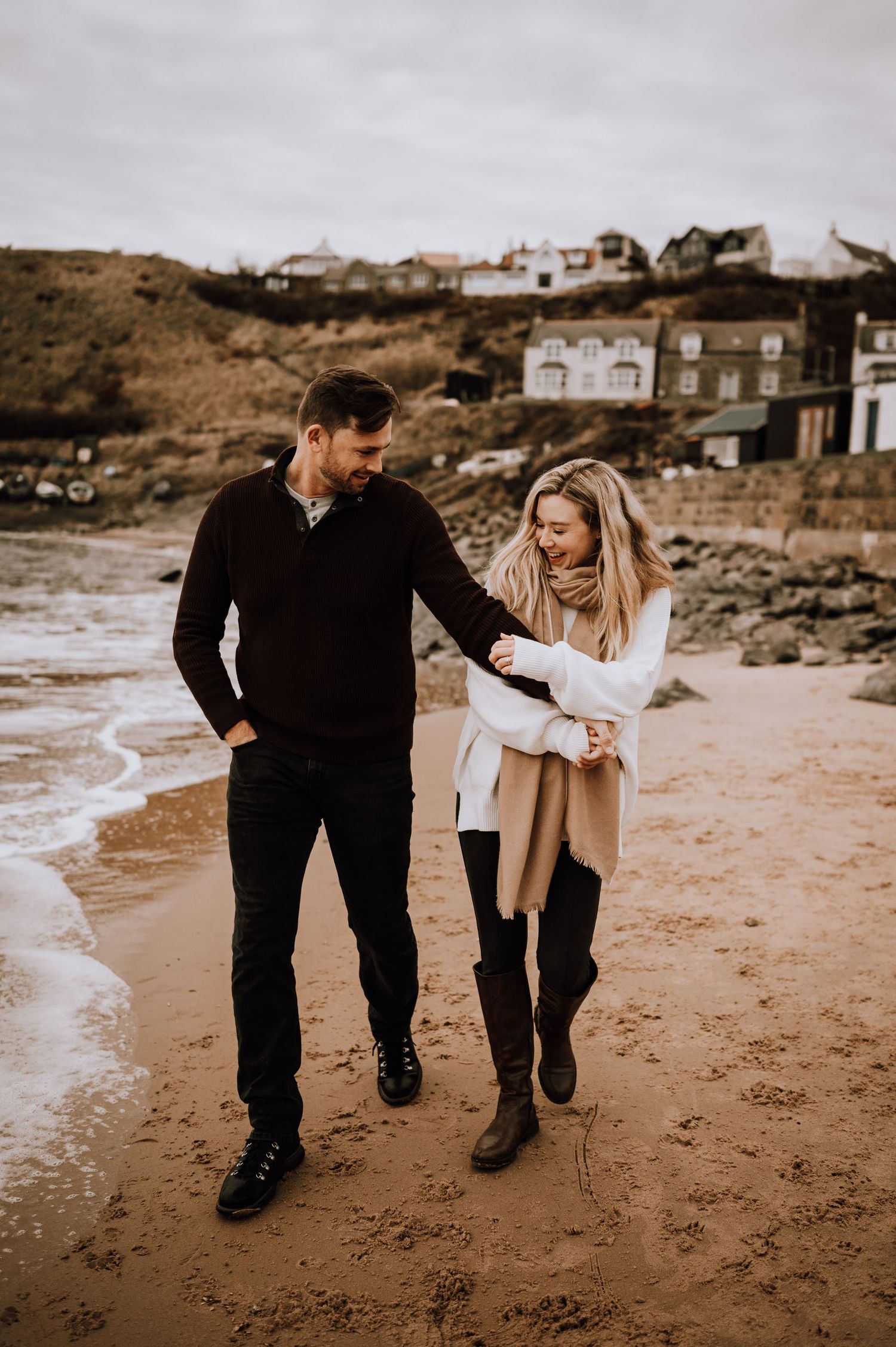 A couple strolling along a scenic beach with coastal homes on the hillside in warm autumn tones.