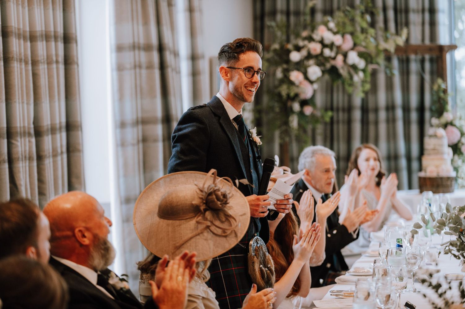 A speaker gives a toast at an elegant wedding reception while guests applaud with glasses raised.
