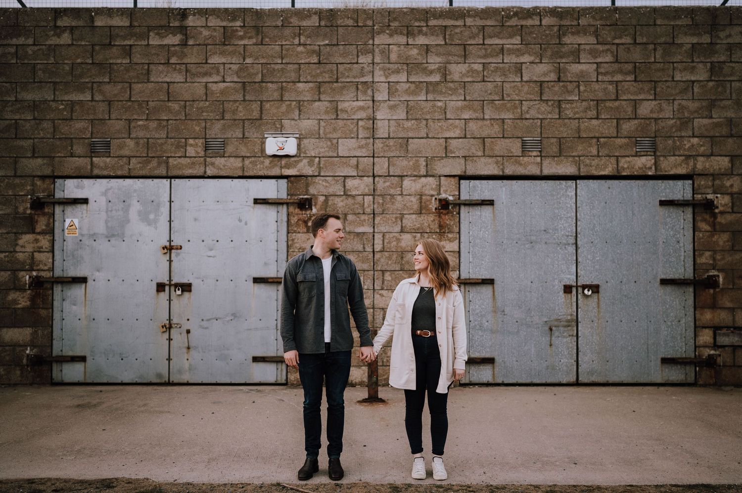 Two people stand against metal doors on a brick wall in an urban setting.
