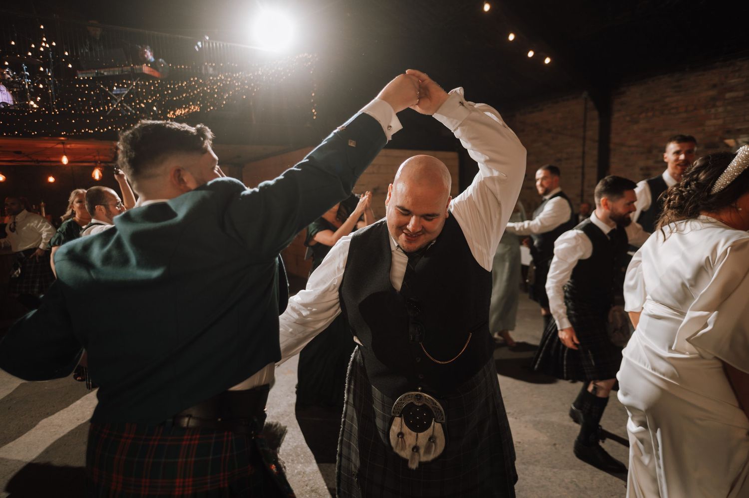 Wedding guests perform traditional Scottish dance moves in kilts at an indoor reception.