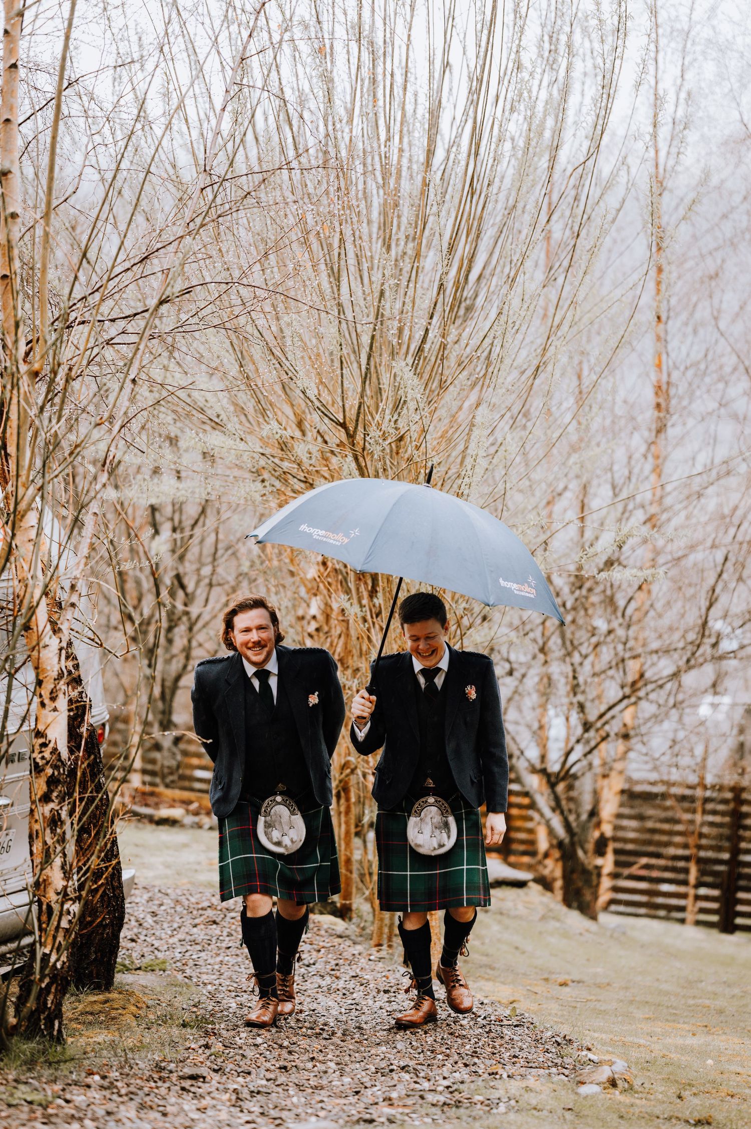 Two people in kilts walk under a gray umbrella among bare winter trees.
