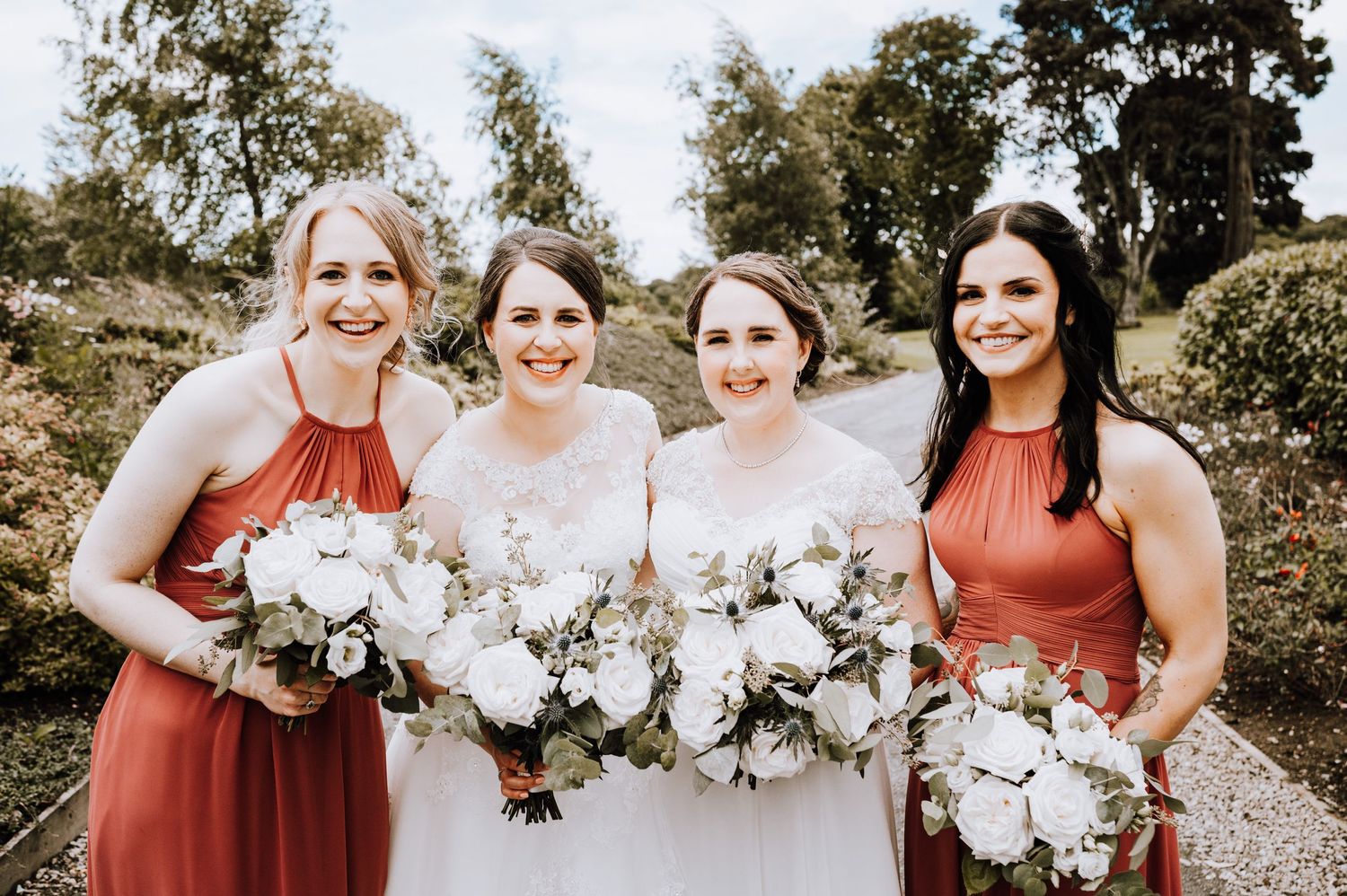 Wedding party poses outdoors with bridesmaids in coral dresses holding white floral bouquets.