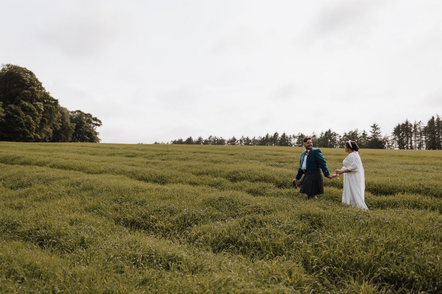 Distant view of wedding couple walking through sunlit meadow in Scottish countryside.