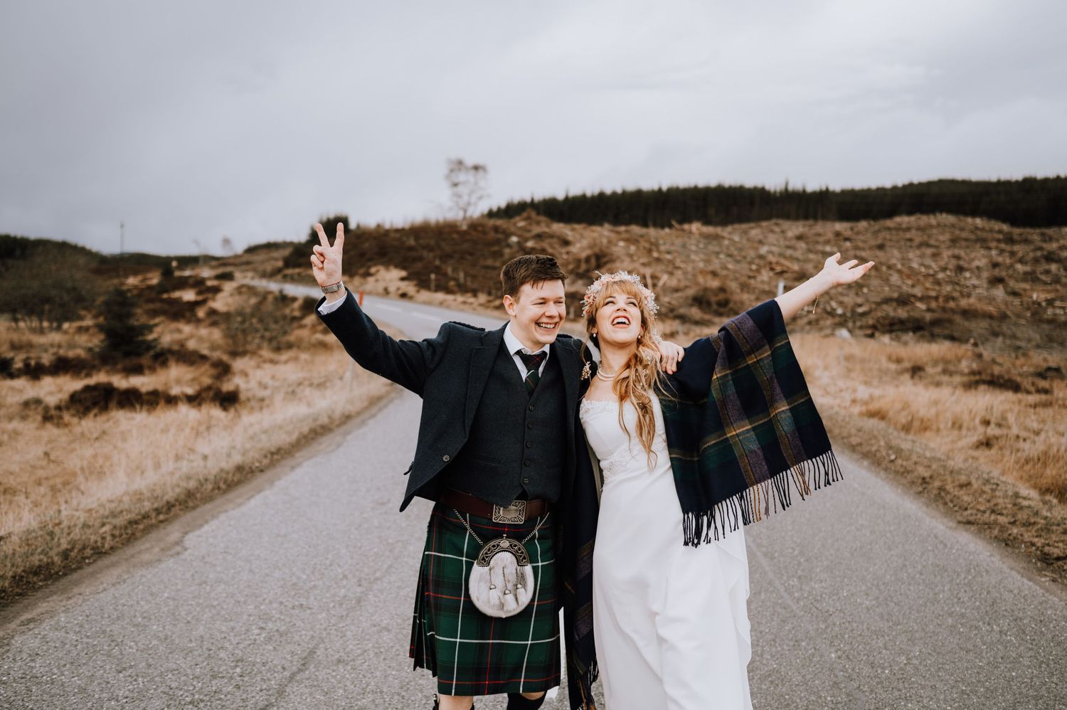 Happy couple raises arms in celebration on Scottish mountain road wearing traditional wedding attire.