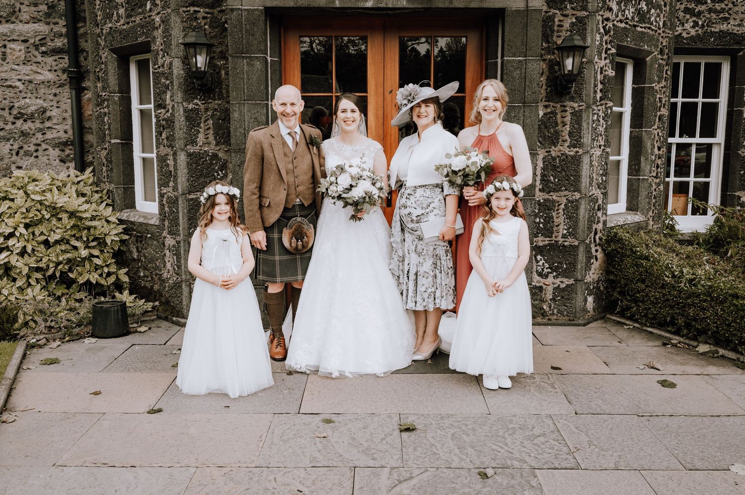 Wedding party group photo outside a Scottish stone building with traditional attire.