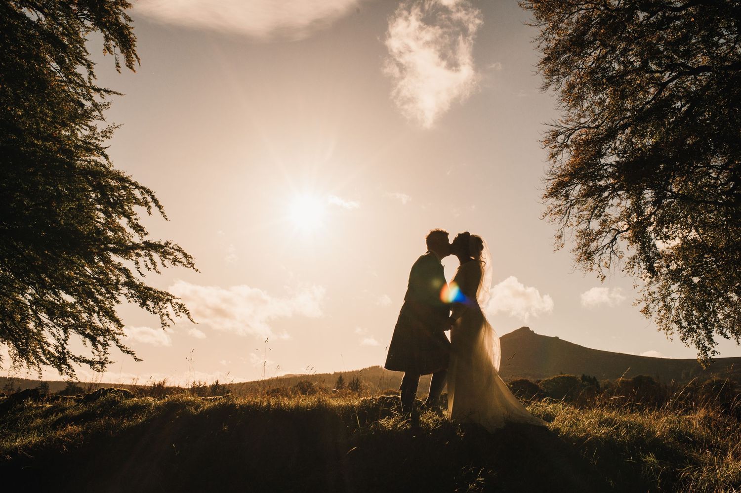 A romantic silhouette of a couple against a sunset sky framed by trees.