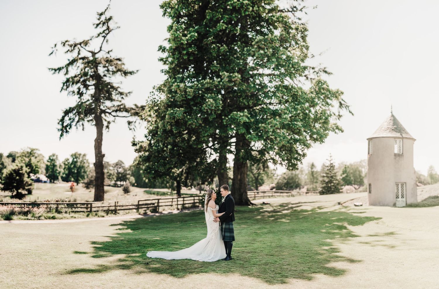 A romantic wedding photo showing a couple embracing in a scenic garden with a white tower visible.