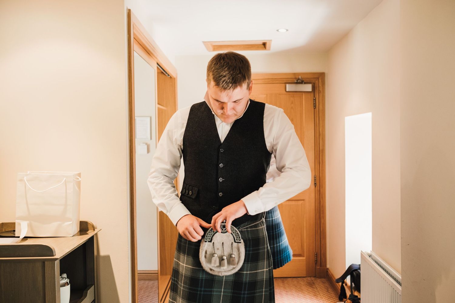 A person in formal Scottish attire adjusts their traditional kilt sporran in an indoor setting.