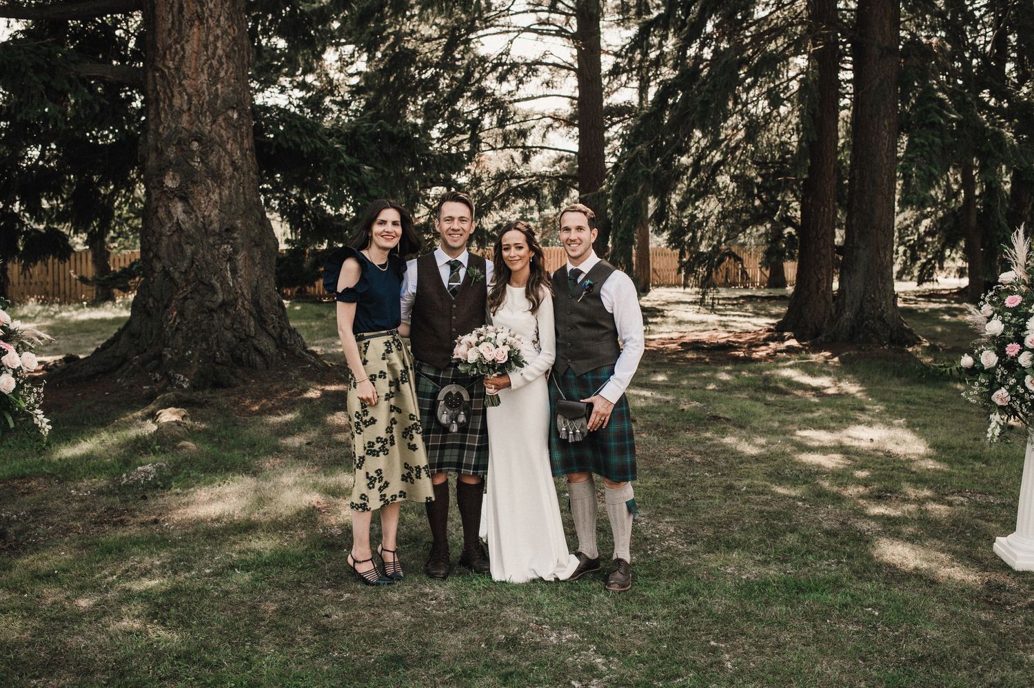 Wedding party members in mix of dresses and kilts pose among tall trees.