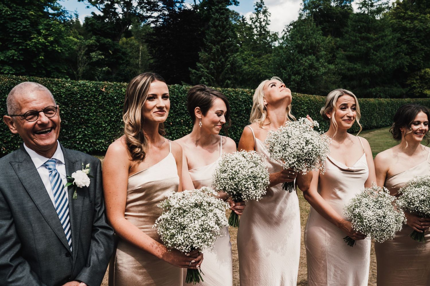 Joyful wedding party laughing together while holding baby's breath bouquets in outdoor garden setting.