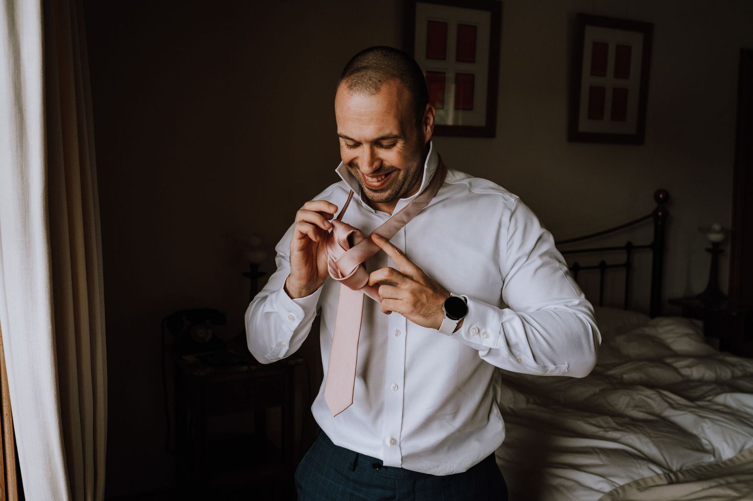 Getting ready scene of adjusting bow tie in bedroom with moody lighting.