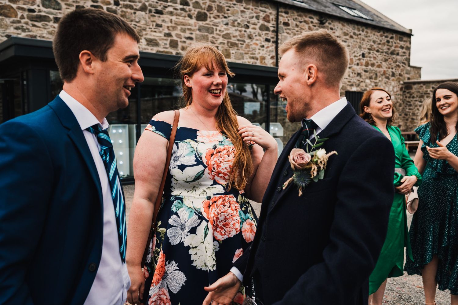 Wedding guests sharing laughter outside a rustic stone building at a Scottish celebration.