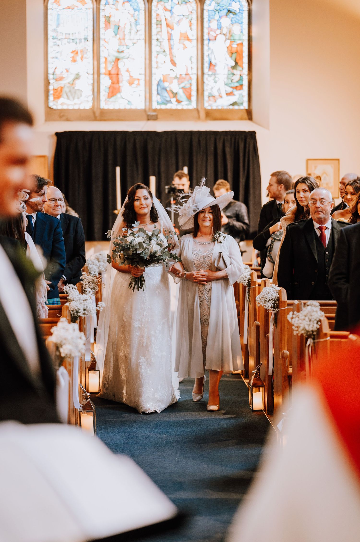 A wedding processional in a church with stained glass windows and lantern-lined aisle.