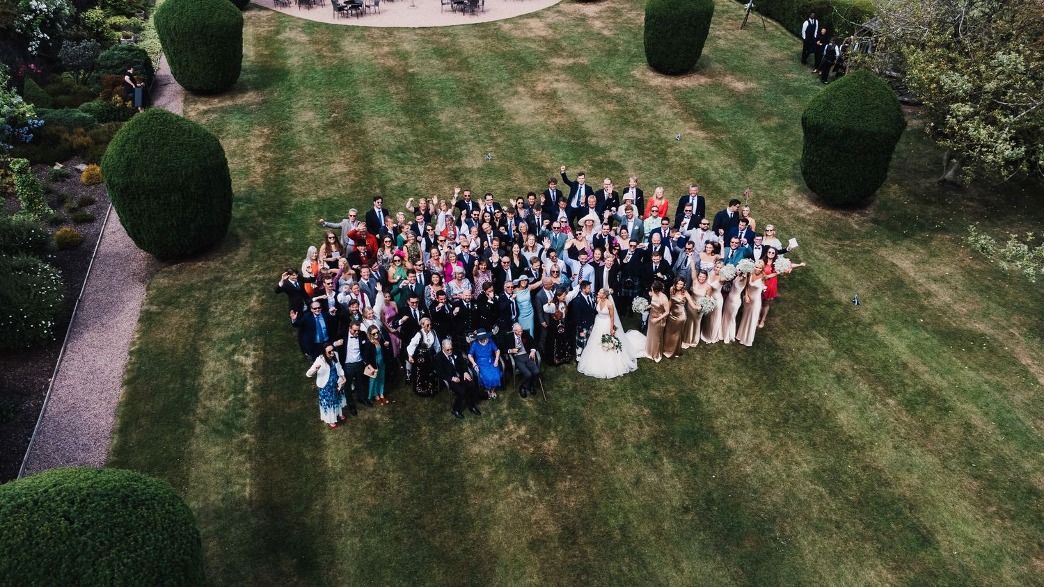 Aerial view of wedding guests gathered on a manicured lawn for a group photo.