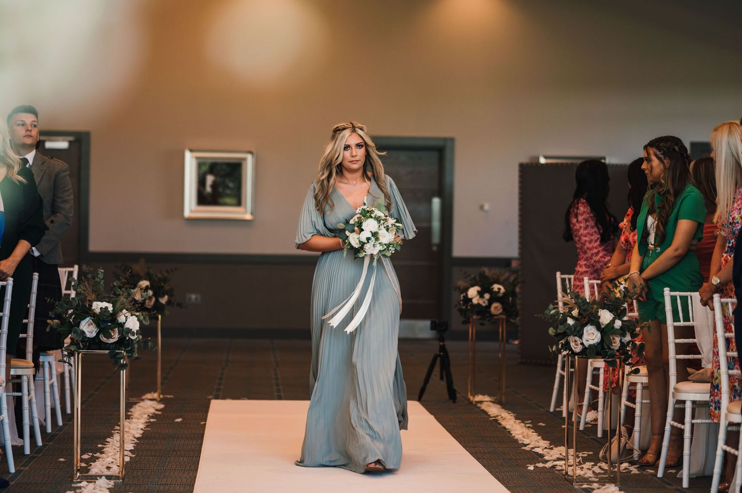Bridesmaid in flowing grey dress walks down aisle carrying white floral bouquet.