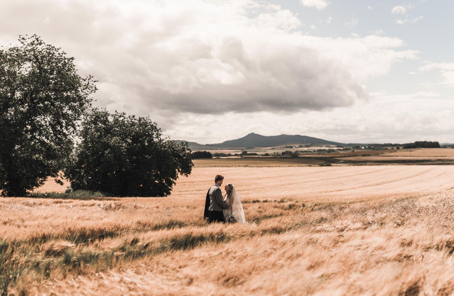 A couple stands in a golden wheat field overlooking distant mountains on a cloudy day.
