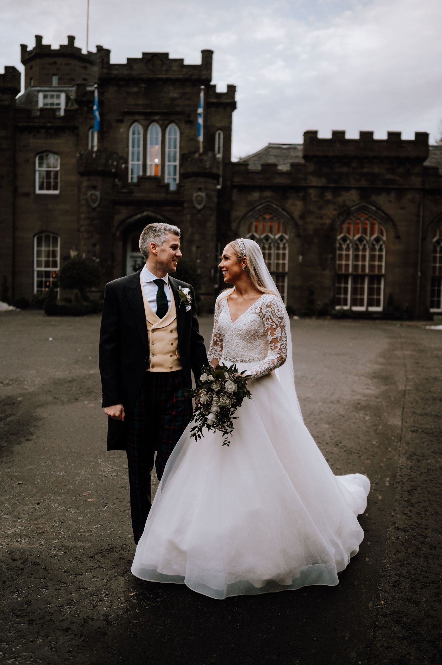 A wedding couple poses in front of a grand gothic castle with stone walls and arched windows.