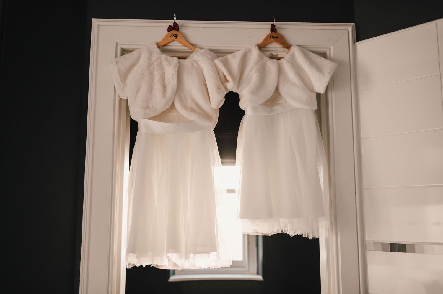Two white wedding dresses hang on wooden hangers against a white door in natural light.