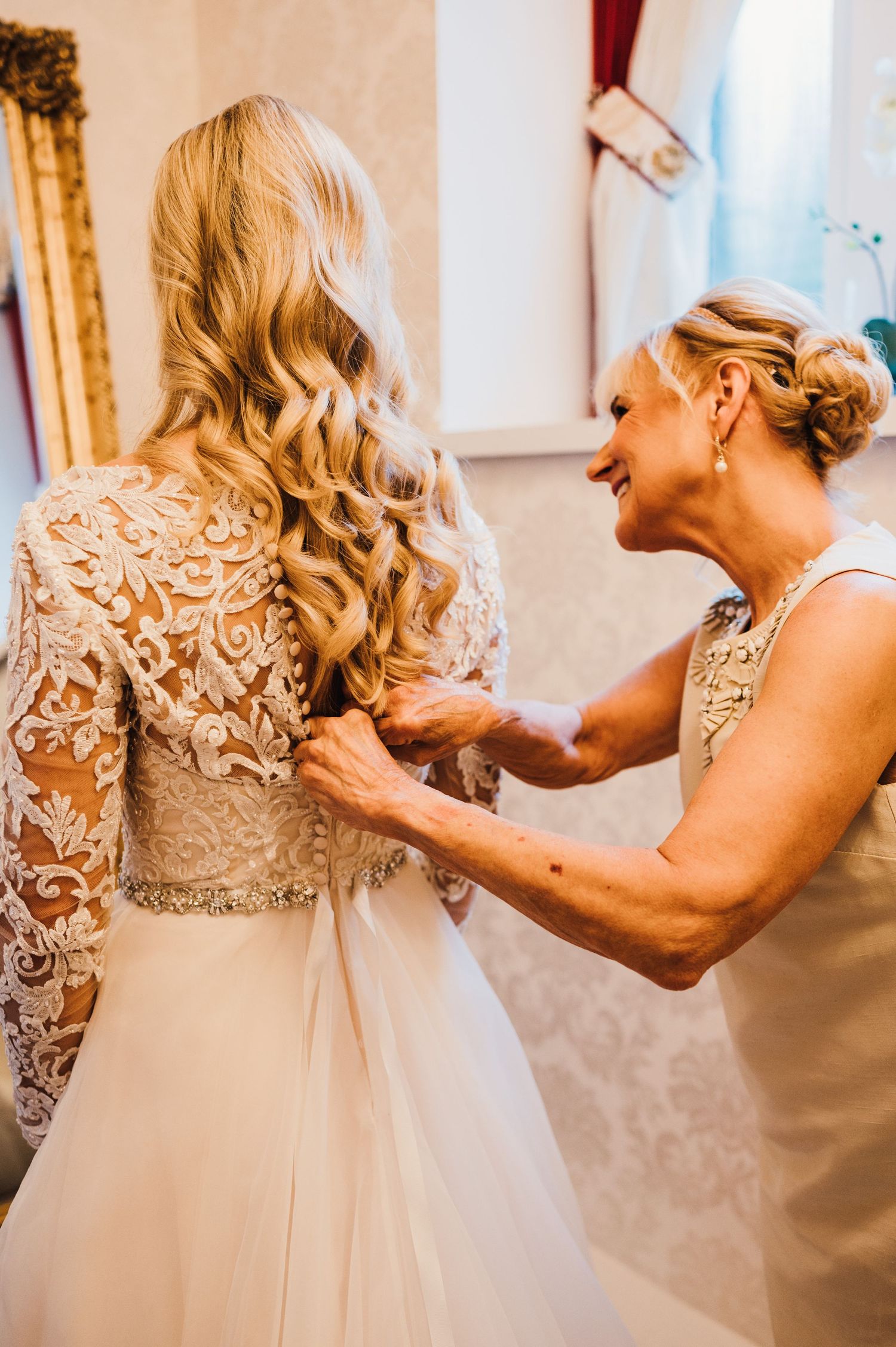 Wedding dress detail being adjusted in elegant room with gold mirror.