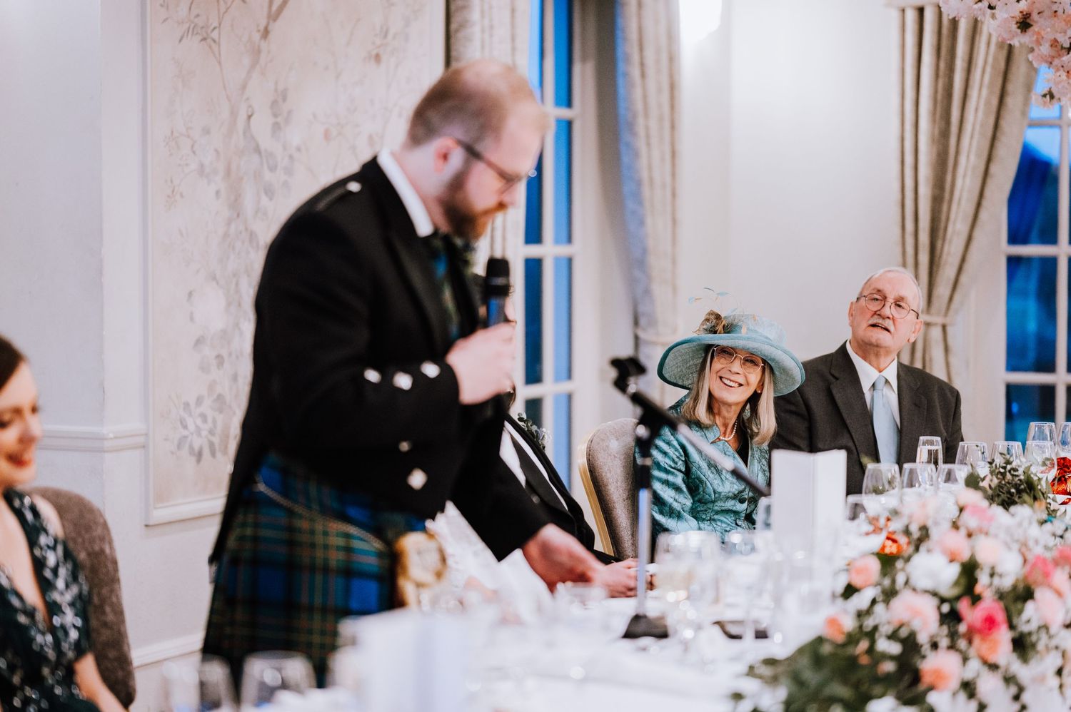 Wedding reception speech at elegant venue with traditional Scottish kilt and formal dining table decor.