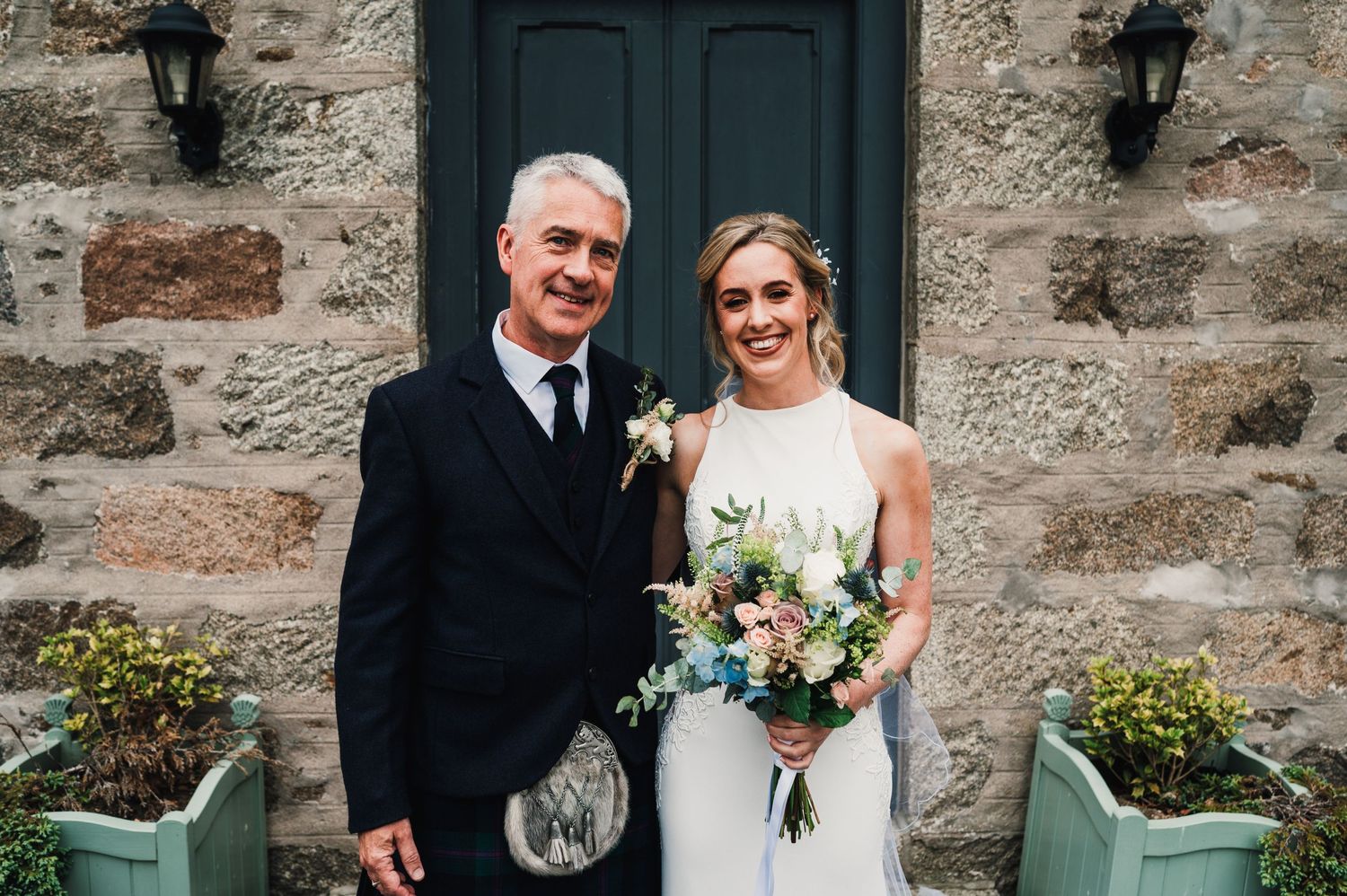 A couple poses outside a stone building with black door wearing wedding attire and traditional Scottish kilt.