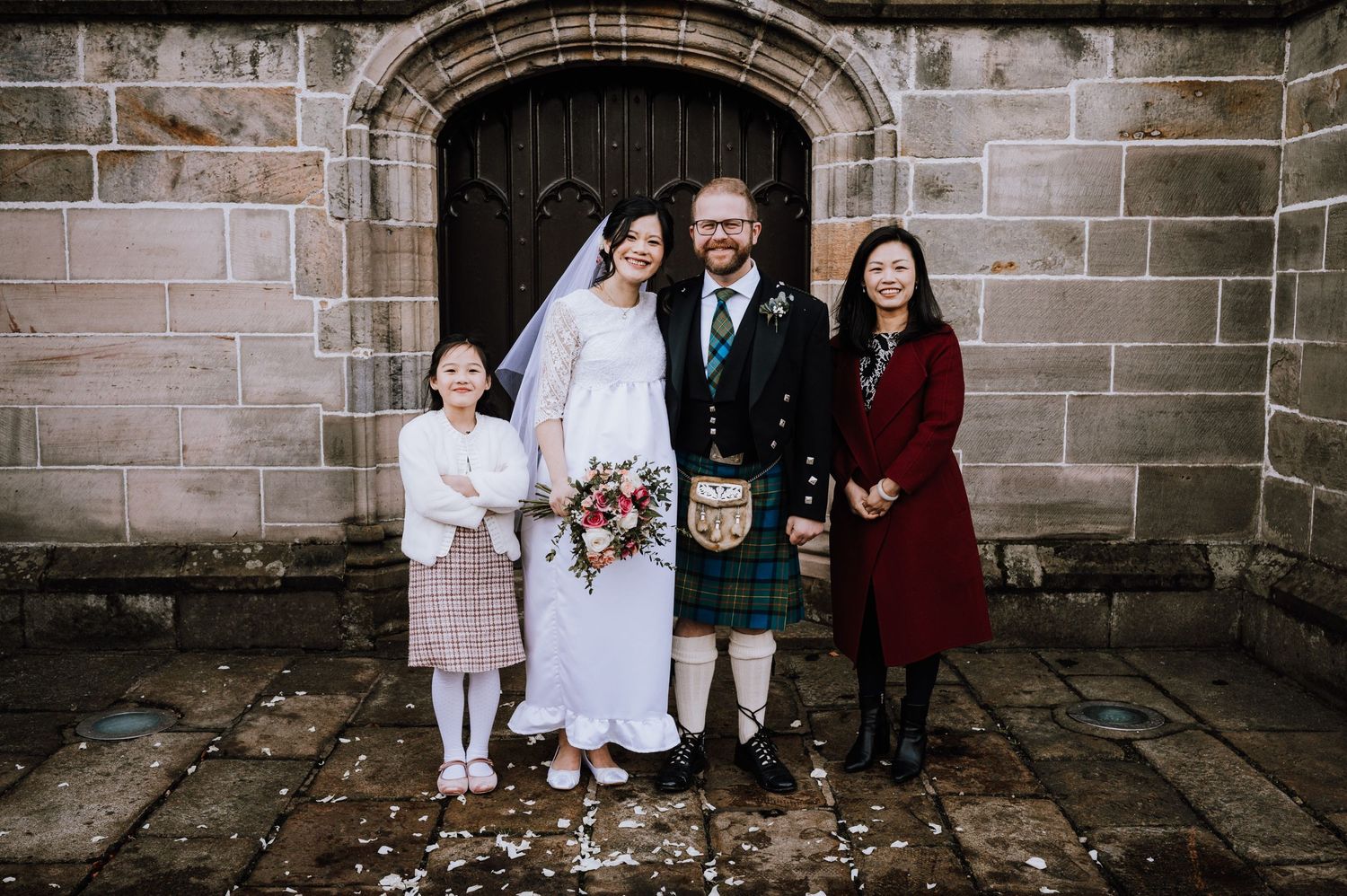 Small wedding party gathering near stone church entrance with Scottish highland dress and formal attire.
