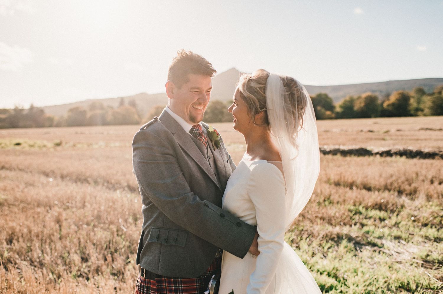 Wedding couple in traditional Scottish attire share intimate moment in sunlit meadow with mountains.
