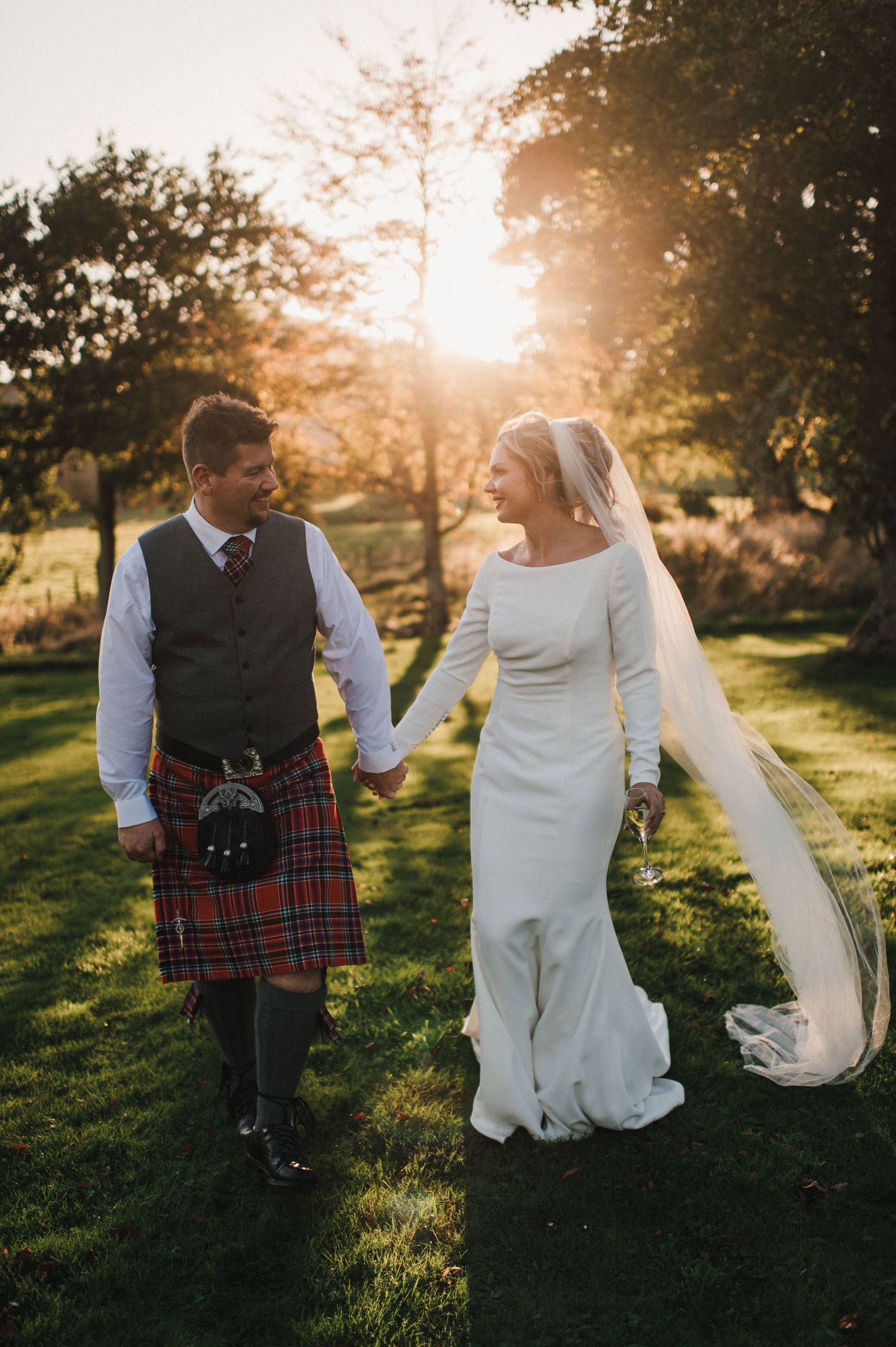 Couple walks hand in hand at sunset, bride in elegant dress and groom wearing traditional Scottish kilt.