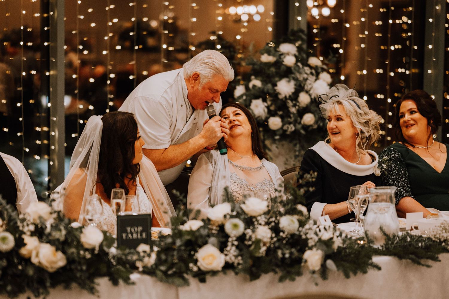 Joyful celebration moment at wedding reception with elegant floral centerpieces and fairy lights.