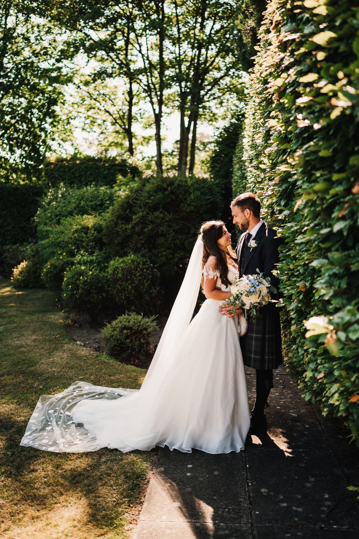 Elegant wedding couple poses on garden path with groom in traditional Scottish kilt and bride in flowing white gown.
