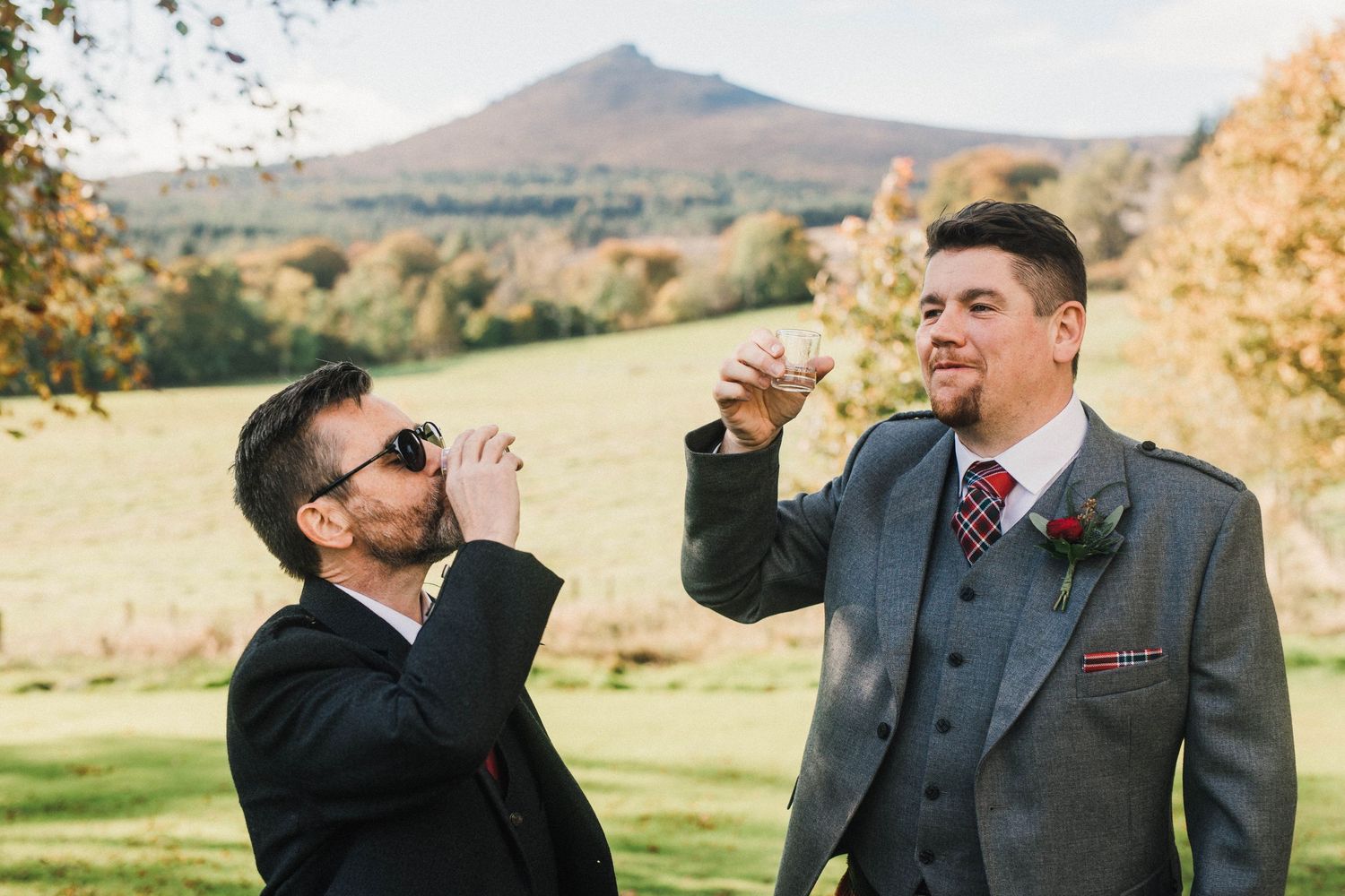 Two people in suits taking celebratory drinks outdoors with mountain in background.