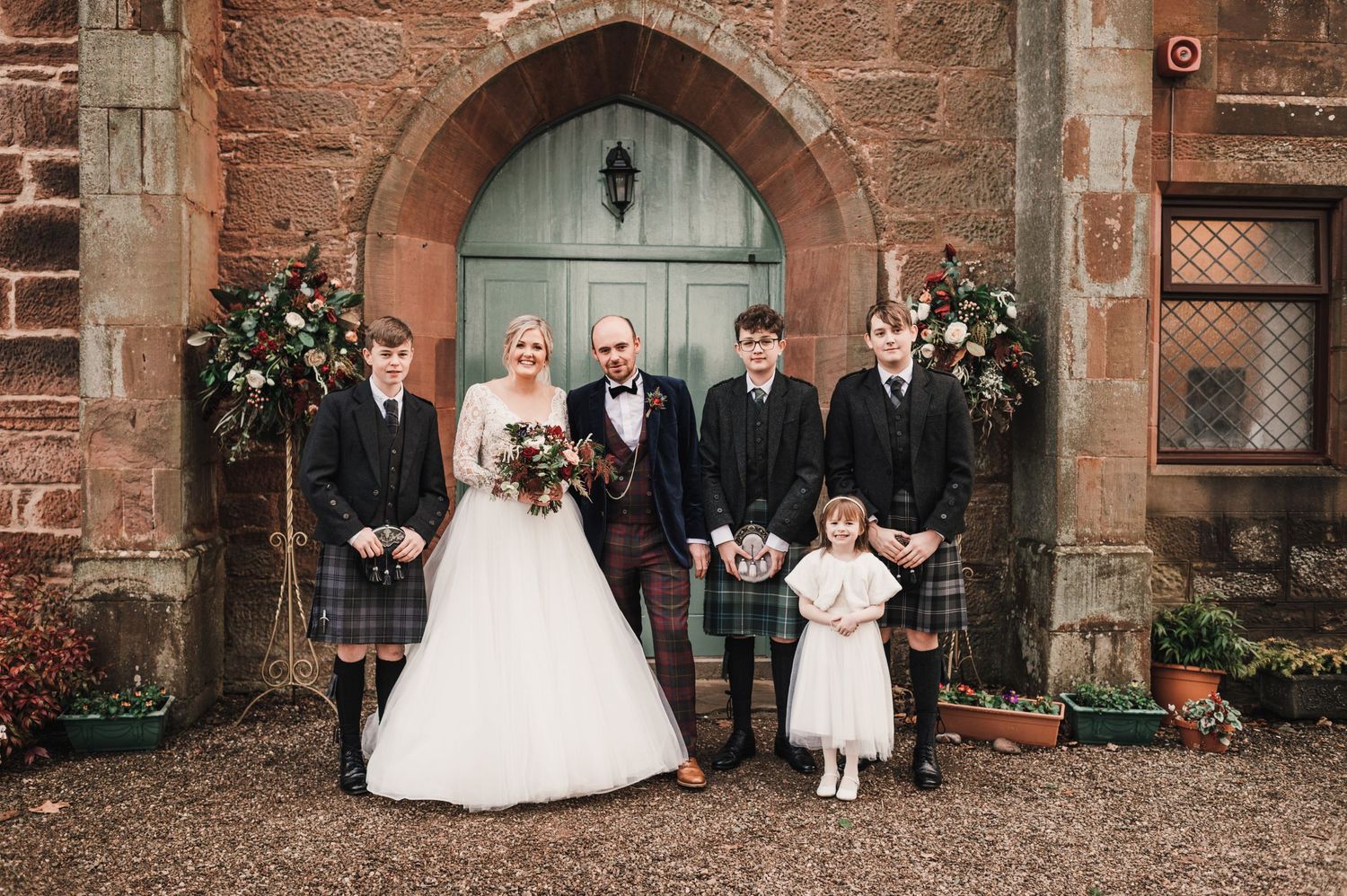 Wedding party members in traditional Scottish attire pose at church entrance.