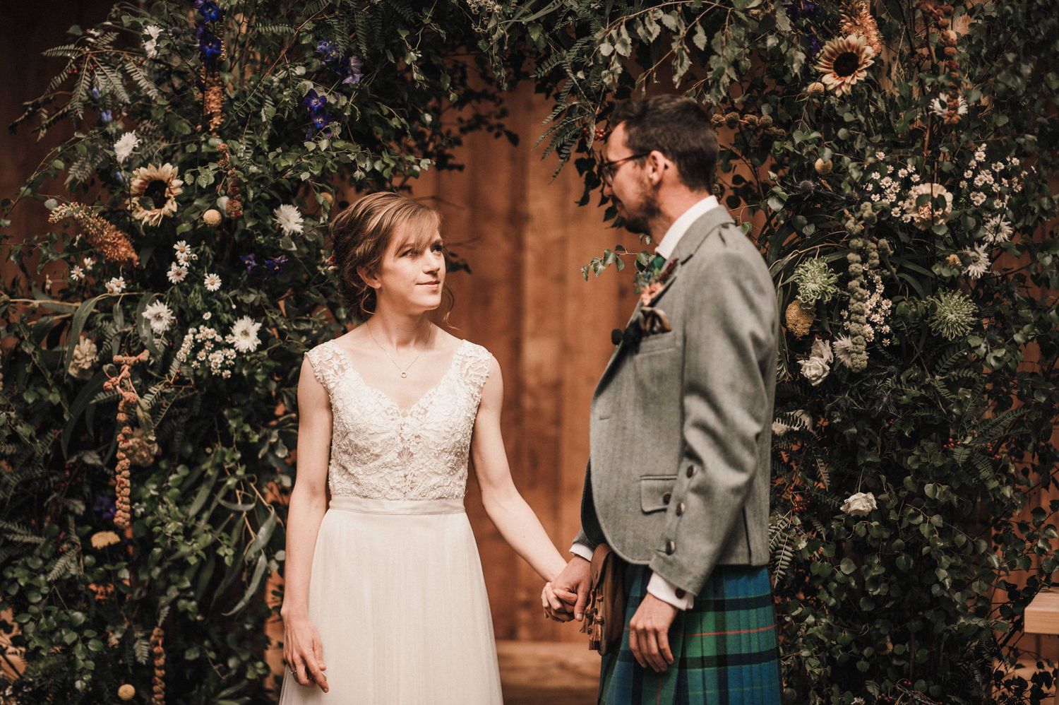 A wedding couple shares a moment under a floral arch with Scottish highland dress elements.