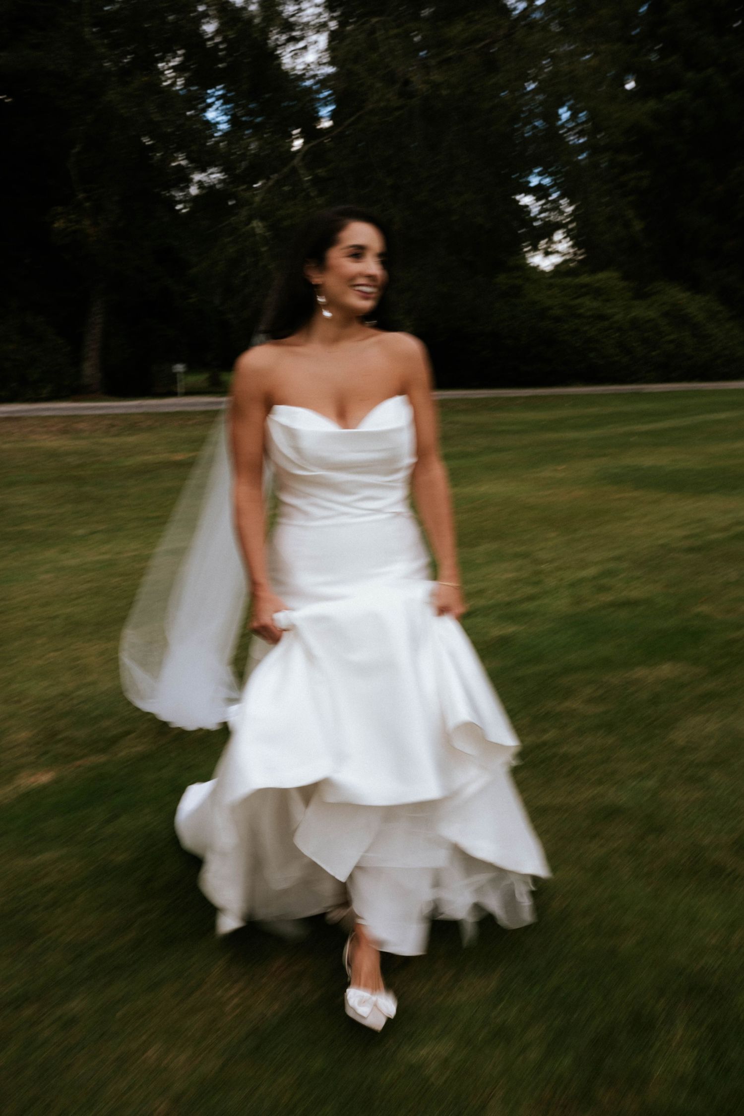 Bride in flowing white dress twirls and dances on green lawn during wedding celebration.
