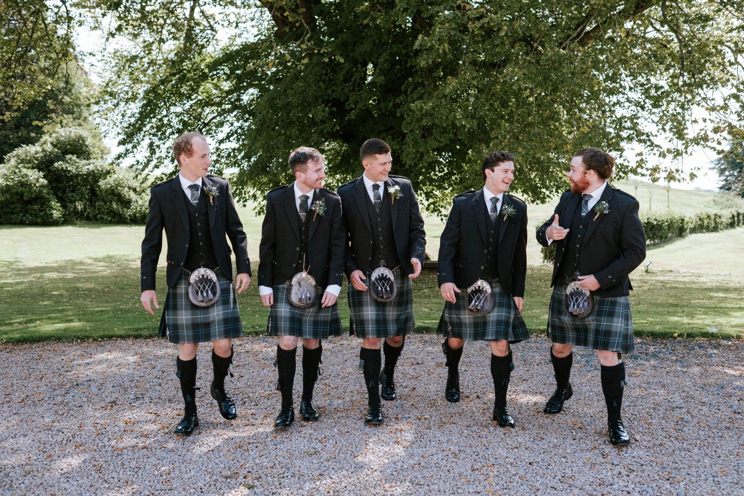 Row of groomsmen in traditional Scottish attire standing outdoors.