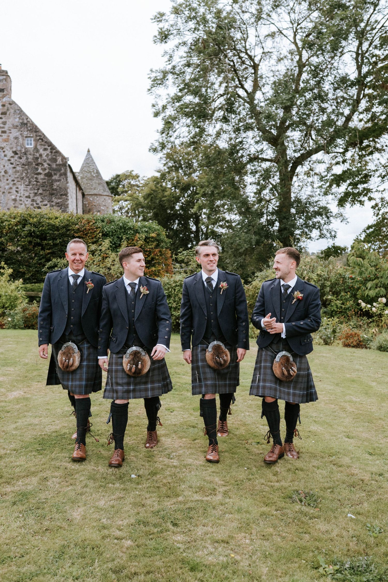 Groomsmen in matching kilts and suits show off their brown dress shoes on grassy hill.