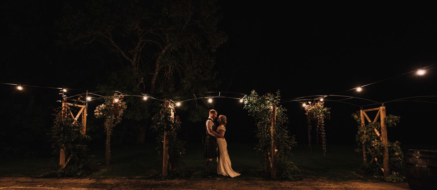 Romantic nighttime wedding ceremony under illuminated wooden arches and string lights.