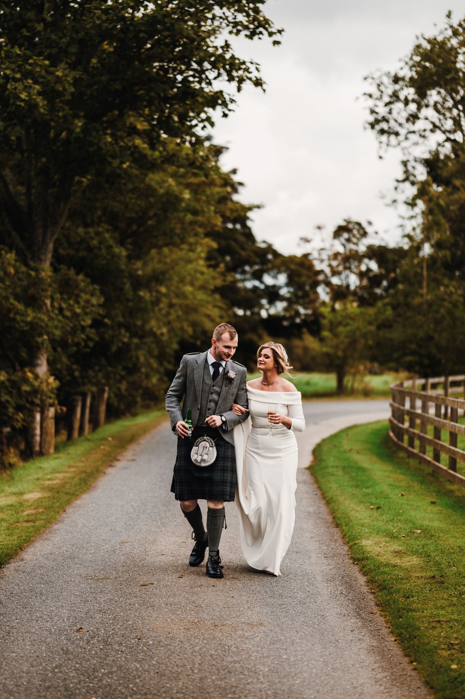 A couple walks along a tree-lined gravel path during a wedding celebration in traditional Scottish attire.