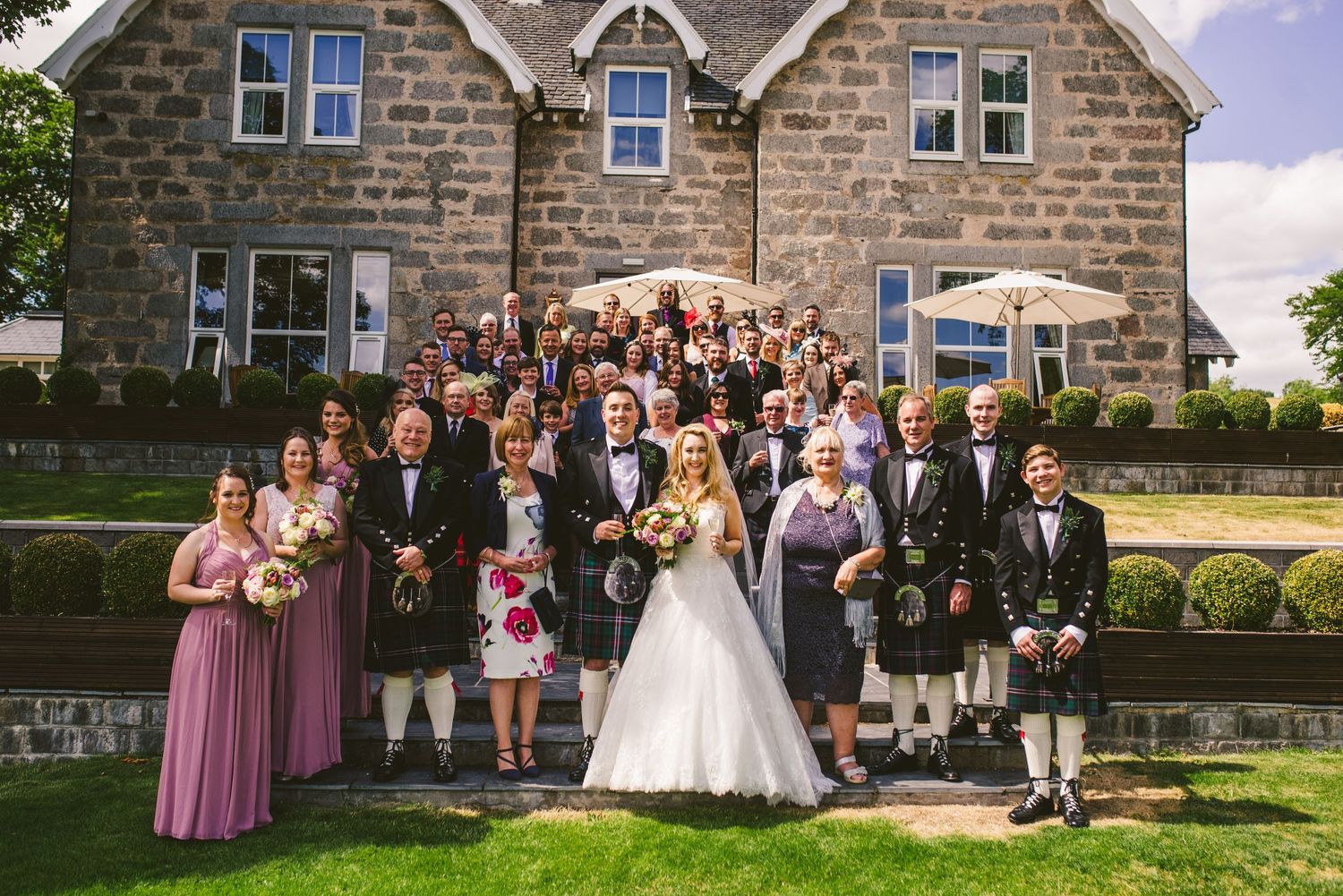 Wedding party poses in front of stone mansion for formal group photos.