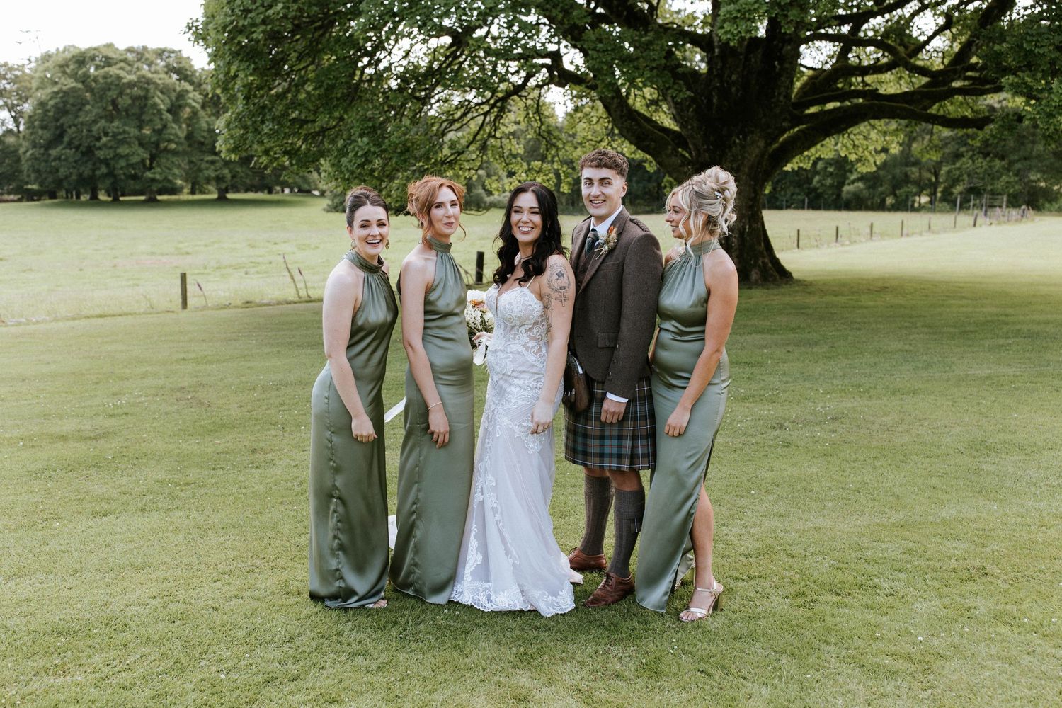 Elegant wedding party poses on green lawn with bridesmaids in sage dresses and traditional Scottish attire.