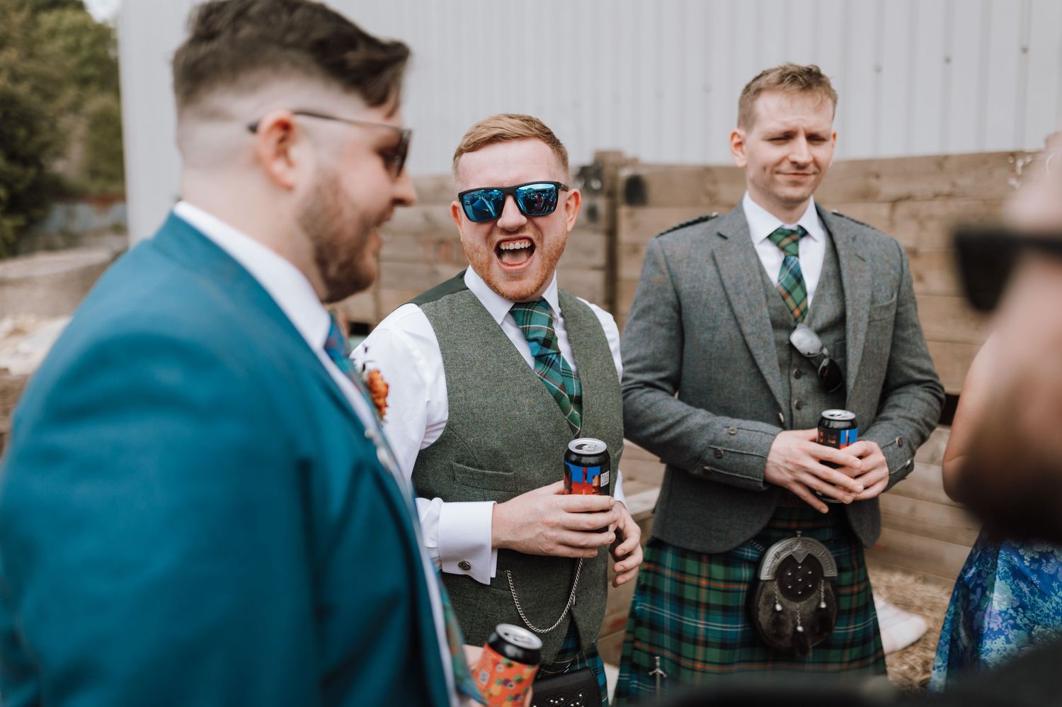 Groomsmen in kilts and suits share drinks during outdoor wedding celebration.
